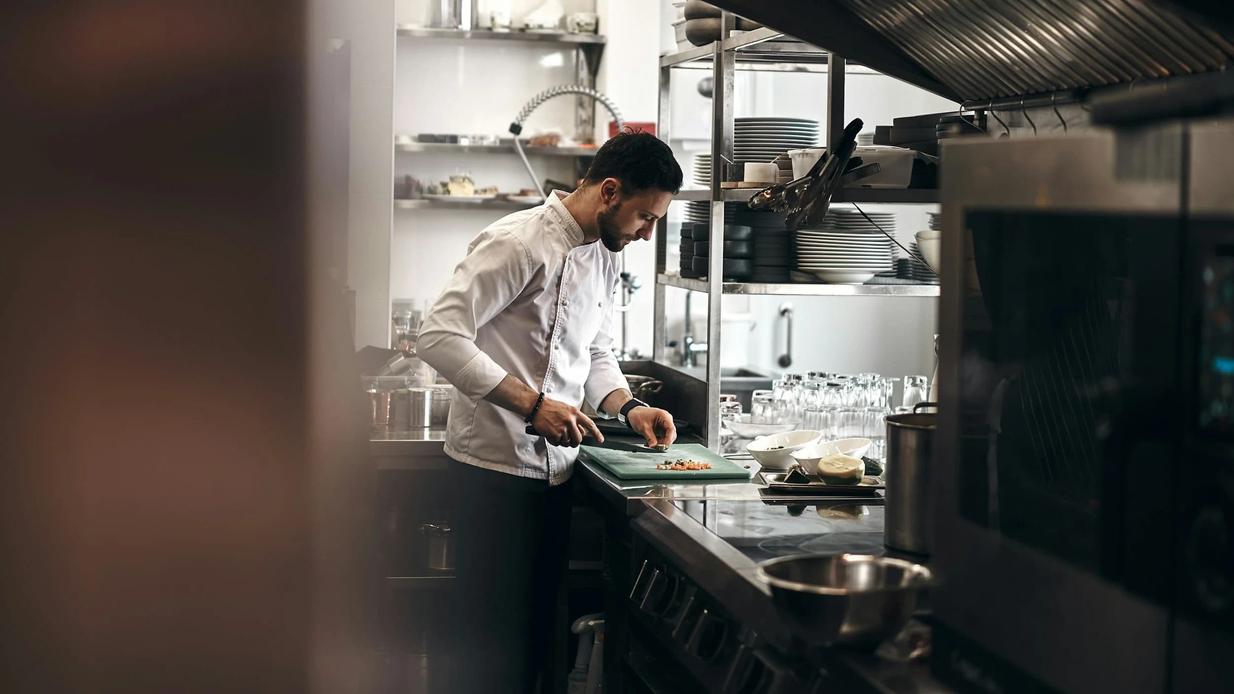 A chef in a white jacket preparing food on a cutting board in a commercial kitchen.