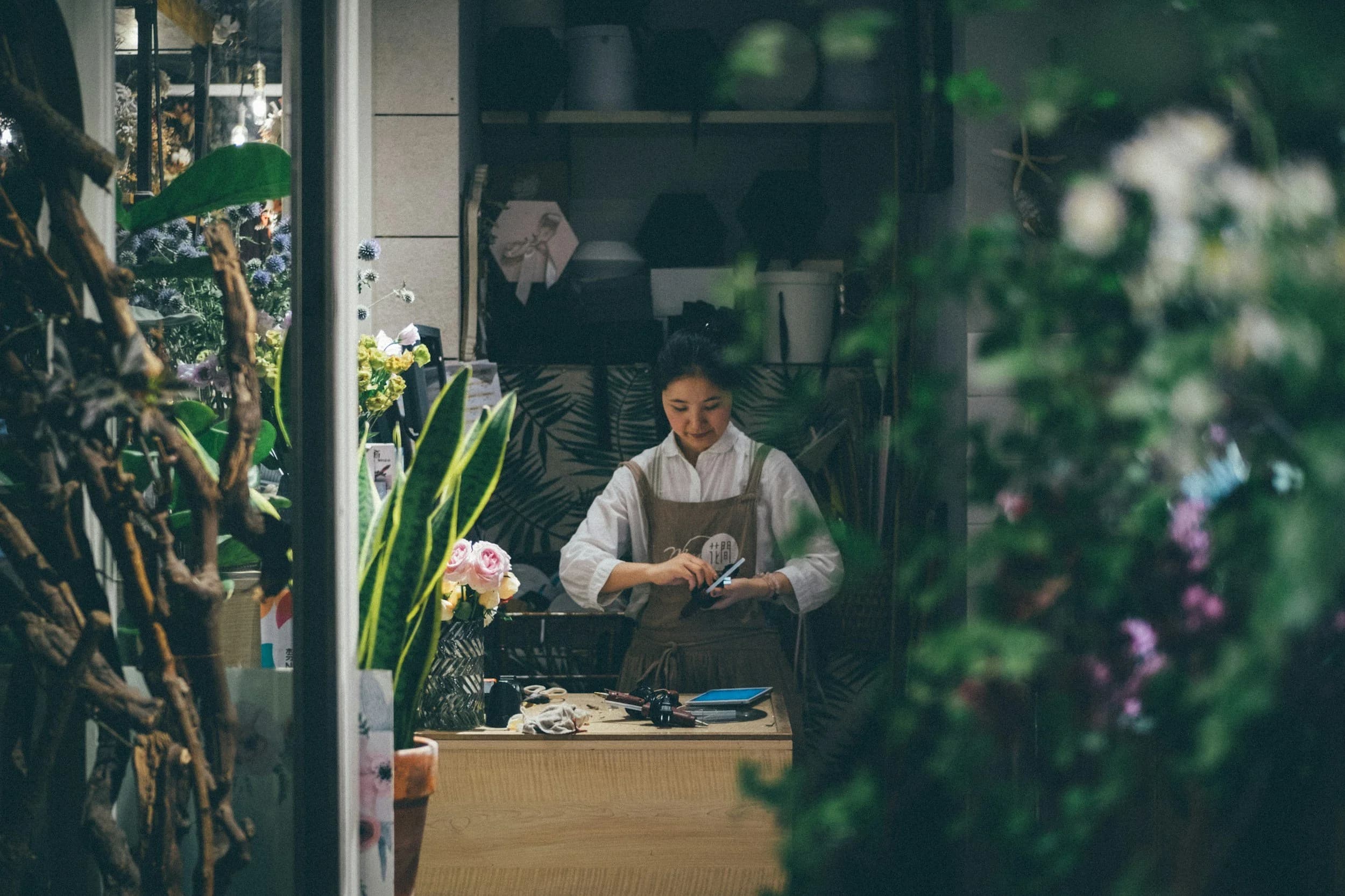 A young woman in a white shirt and apron standing behind a wooden counter inside a flower shop.