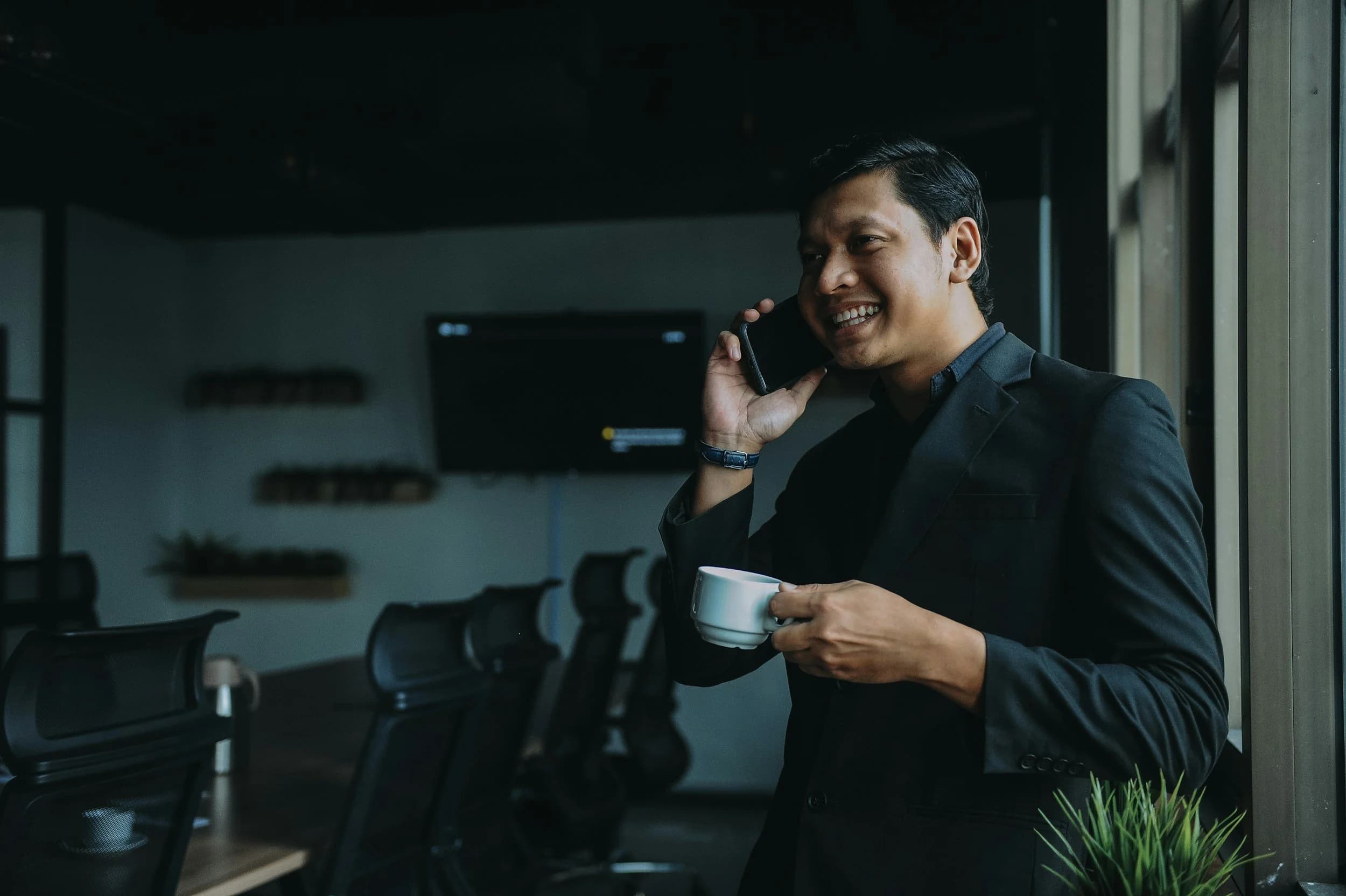 A smiling man in a dark suit talking on a phone in a modern office.